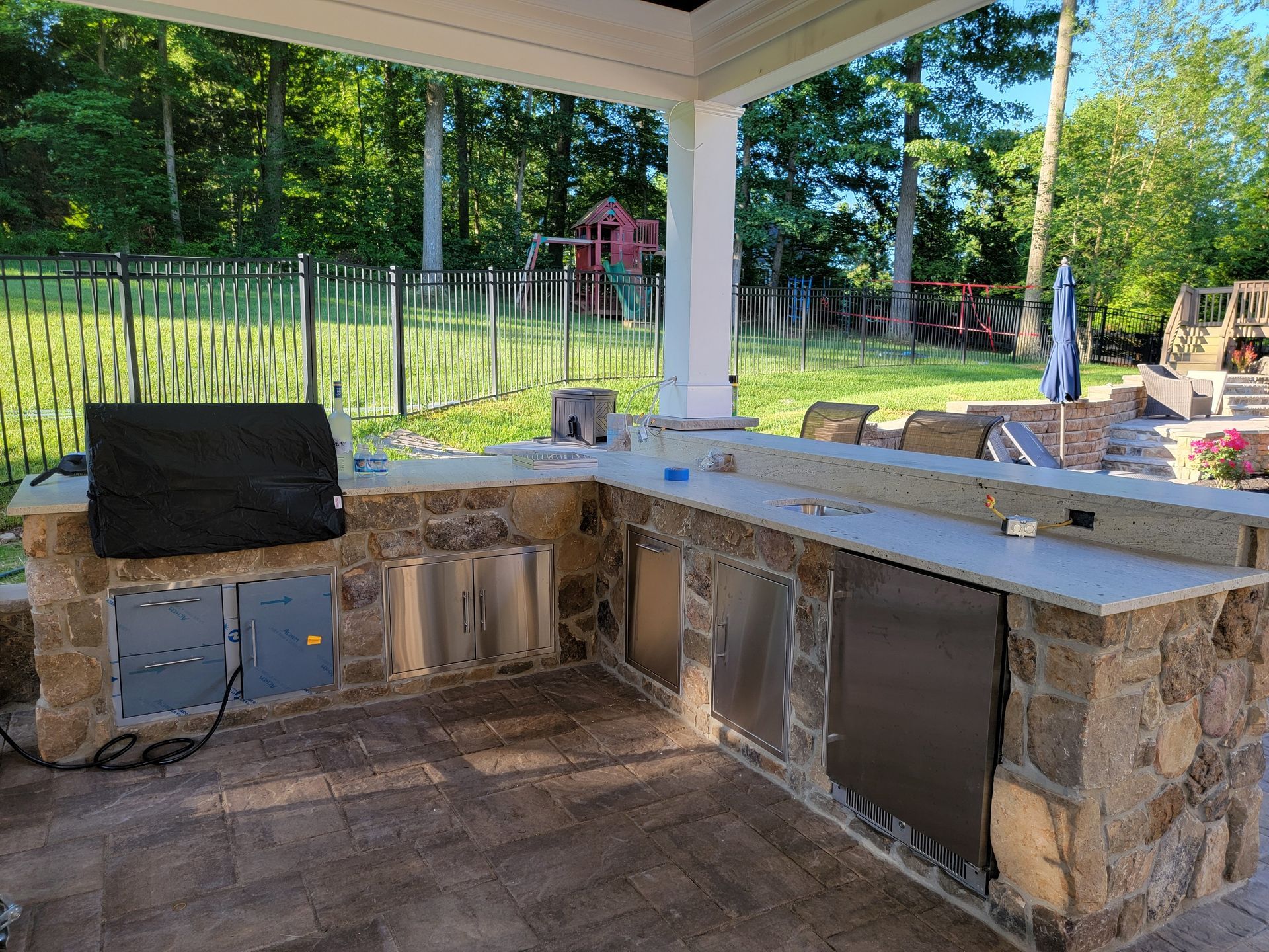Outdoor kitchen with stone counters, stainless steel appliances, under a covered patio.