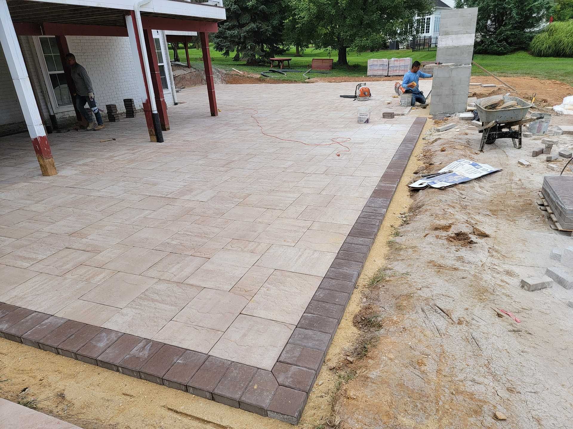 Patio being constructed with tan pavers and a brown border; a worker is in the background.