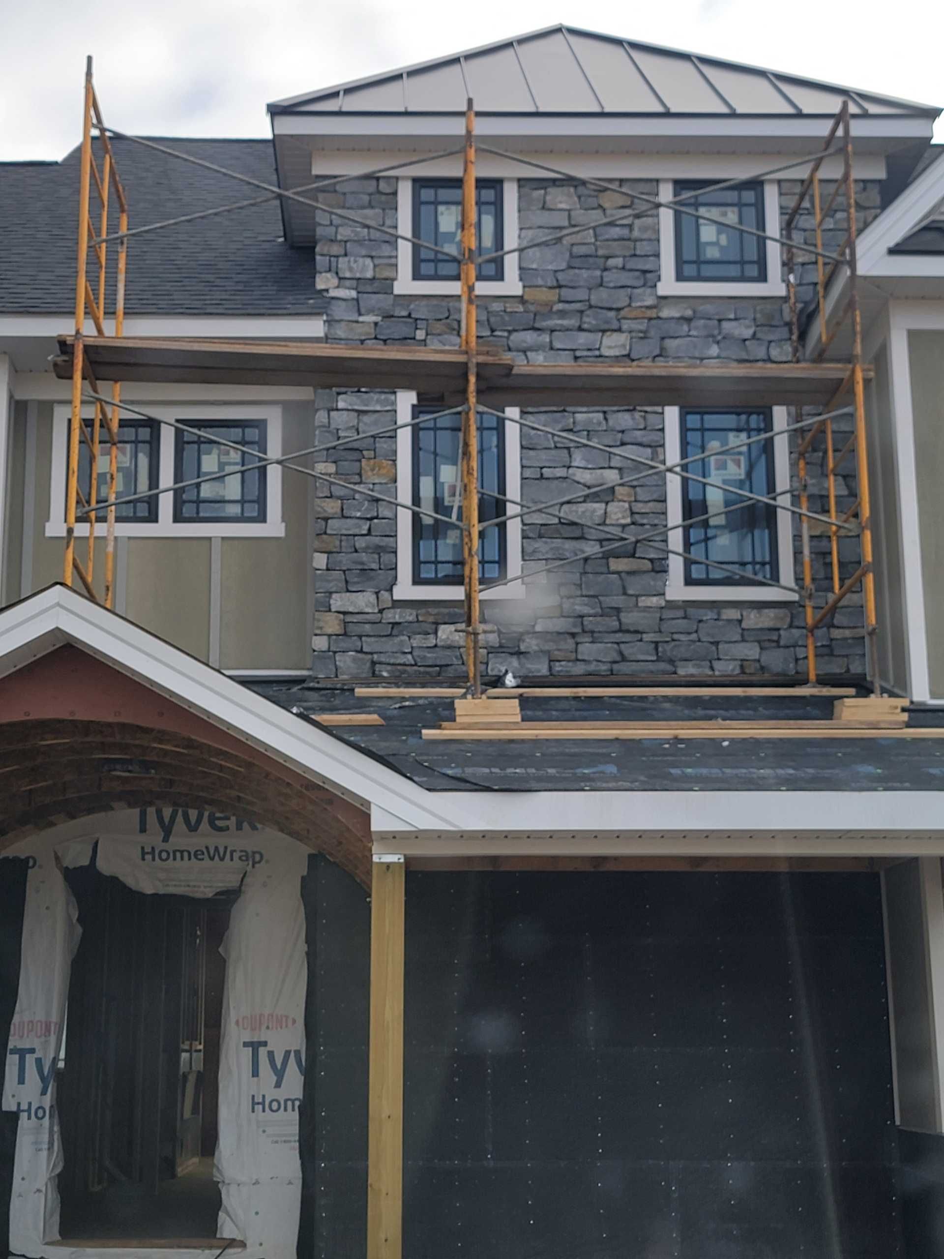 House exterior under construction, with scaffolding around a stone facade, beige siding, and a dark garage door.