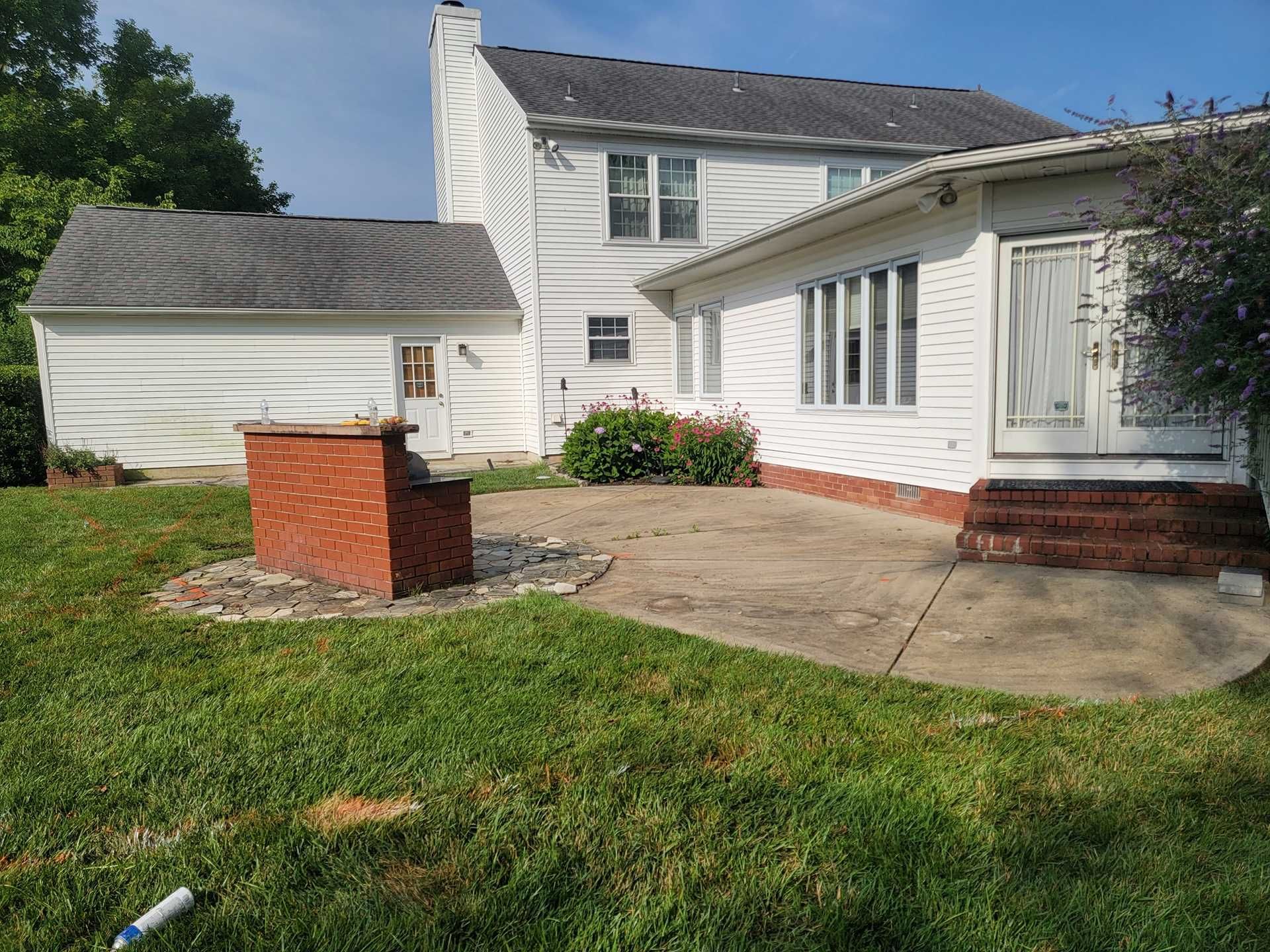 Backyard with a brick grill, concrete patio, white house, and green lawn.