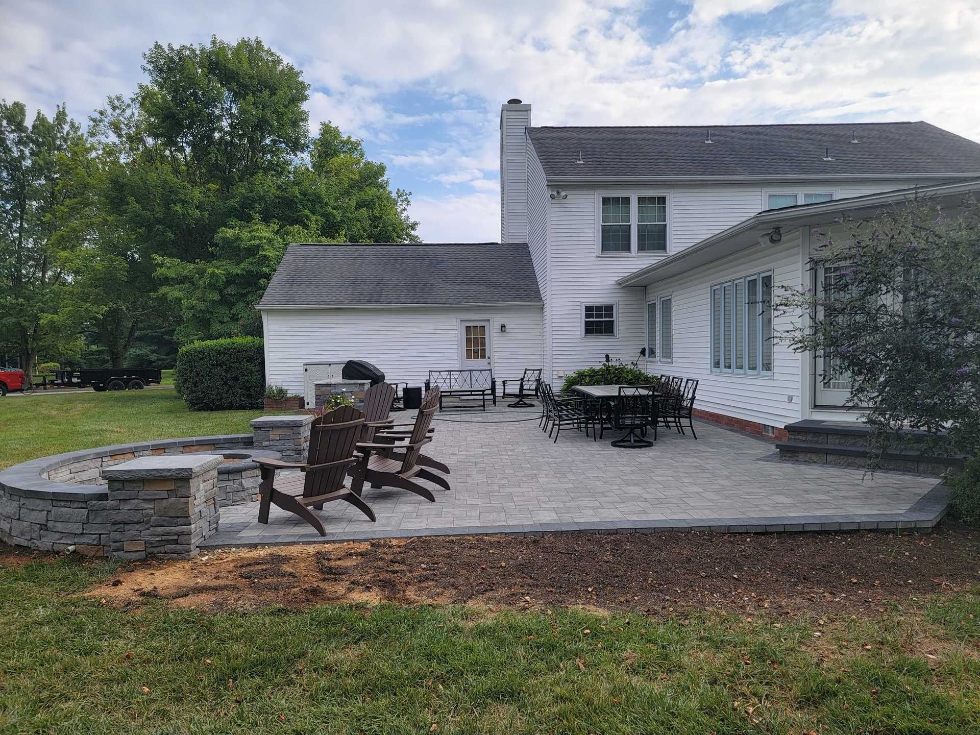 Backyard patio with stone pavers, fire pit, seating, and a white house.
