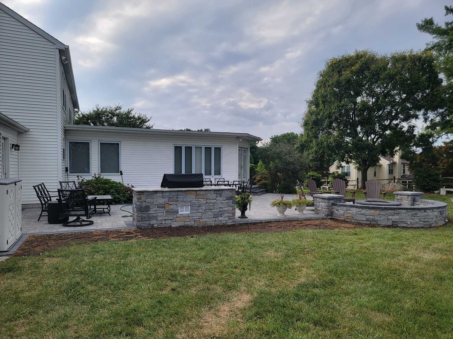 Backyard with stone grill, patio furniture, and flower beds, under a cloudy sky.