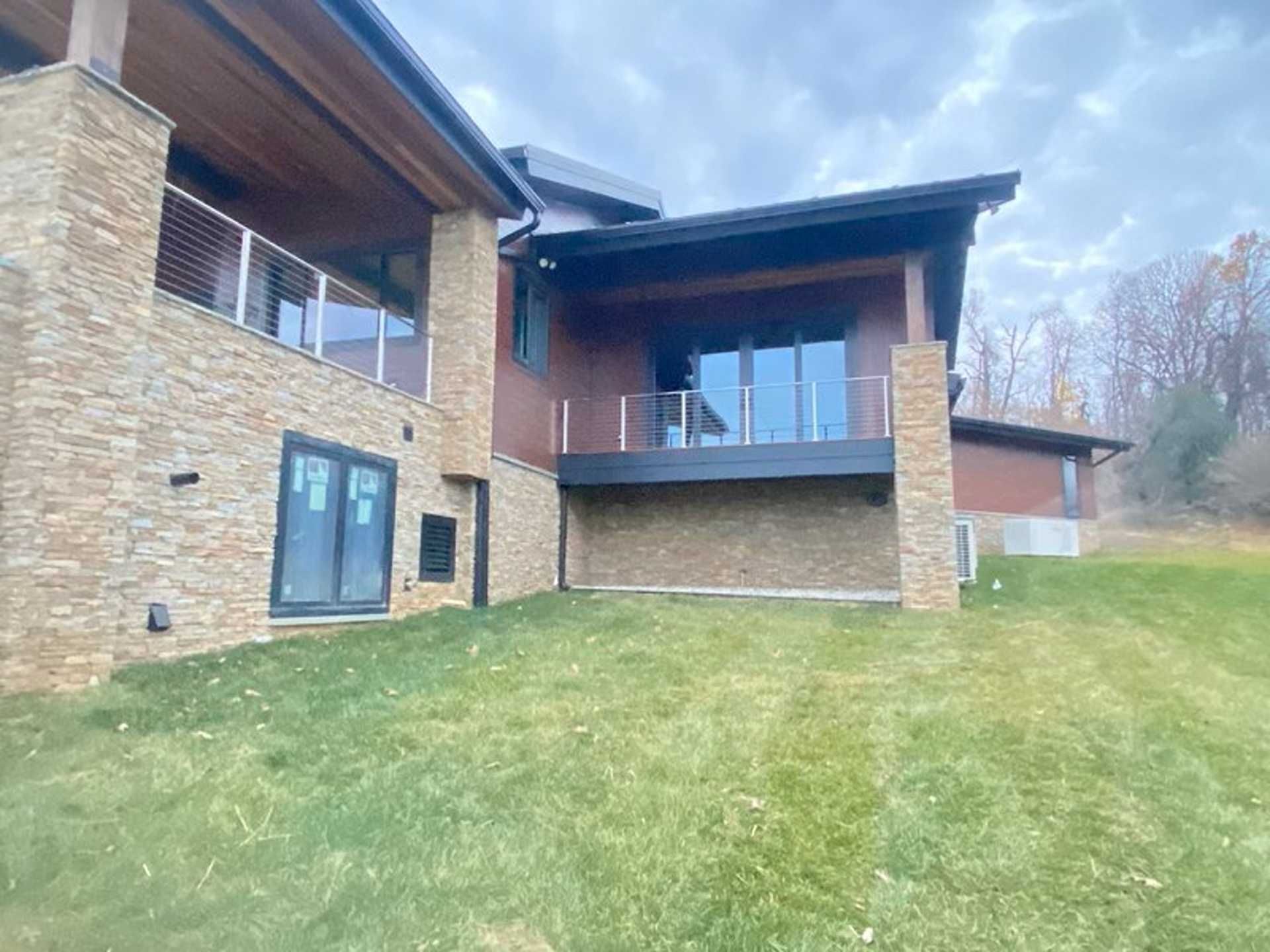 Exterior view of a modern house with stone facade, glass railings, and a grassy lawn under a cloudy sky.