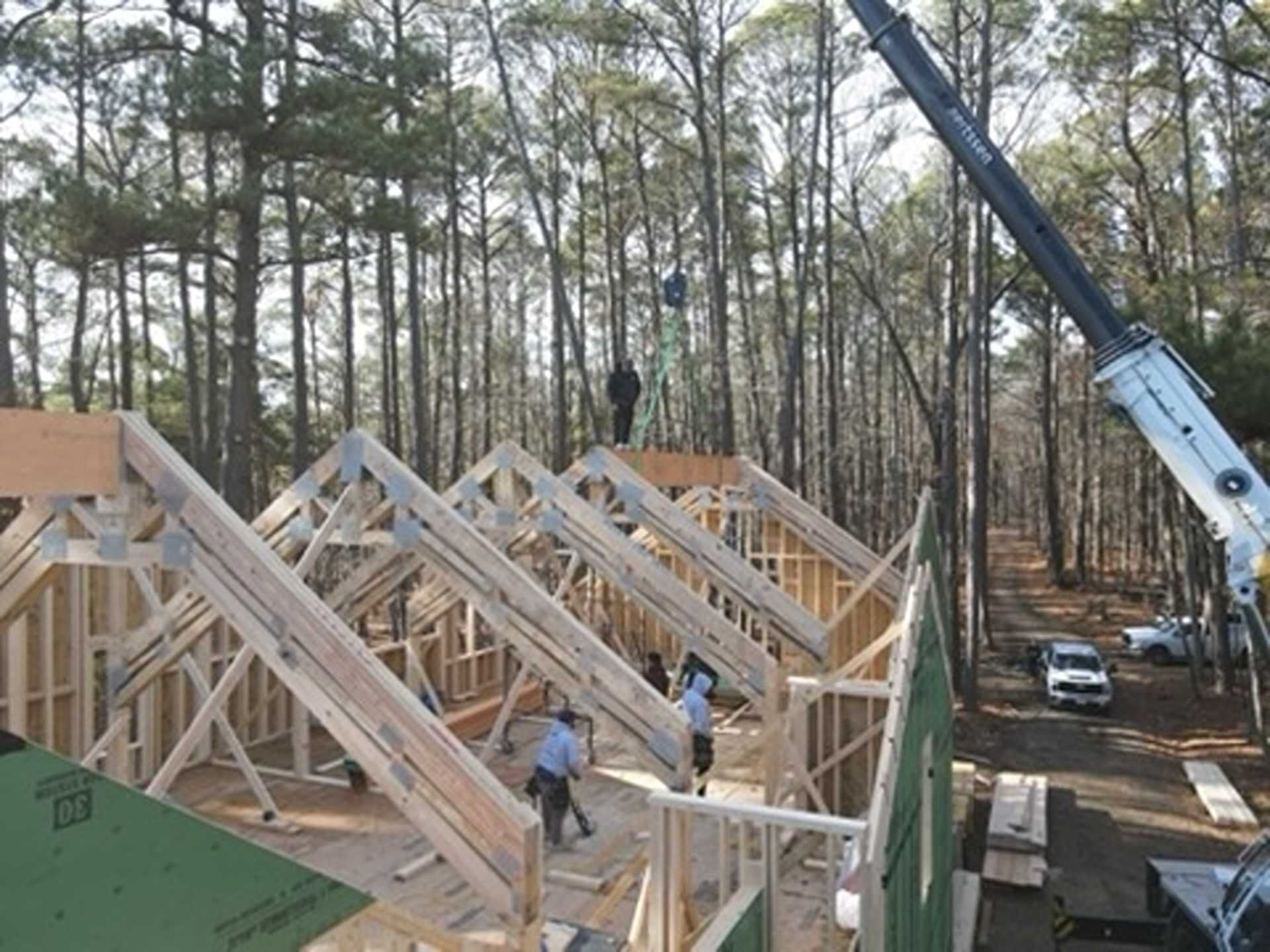 Construction workers installing roof trusses on a house in a wooded area. A crane assists.