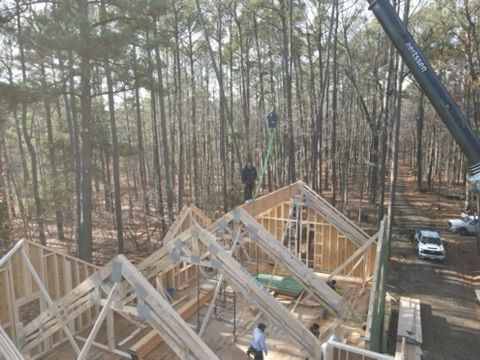 Construction workers framing a house with a crane in a wooded area.
