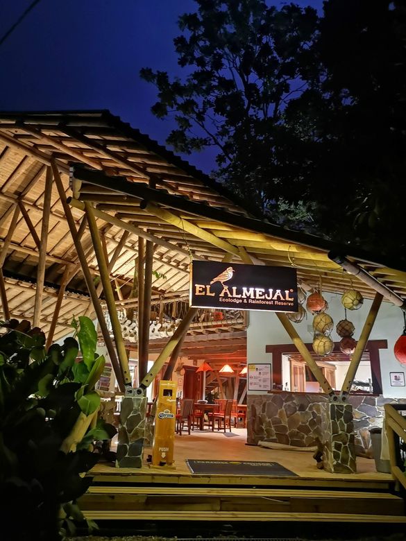 Open-air restaurant at dusk with bamboo roof, warm lights, and a glowing sign amid tropical greenery