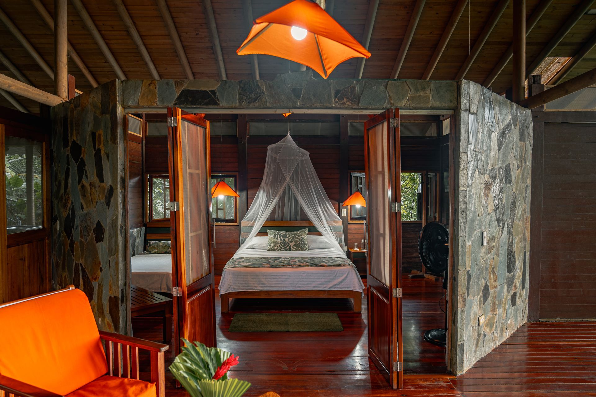 Bedroom interior with an orange chair, stone walls, open wooden doors, and a bed under a mosquito net.