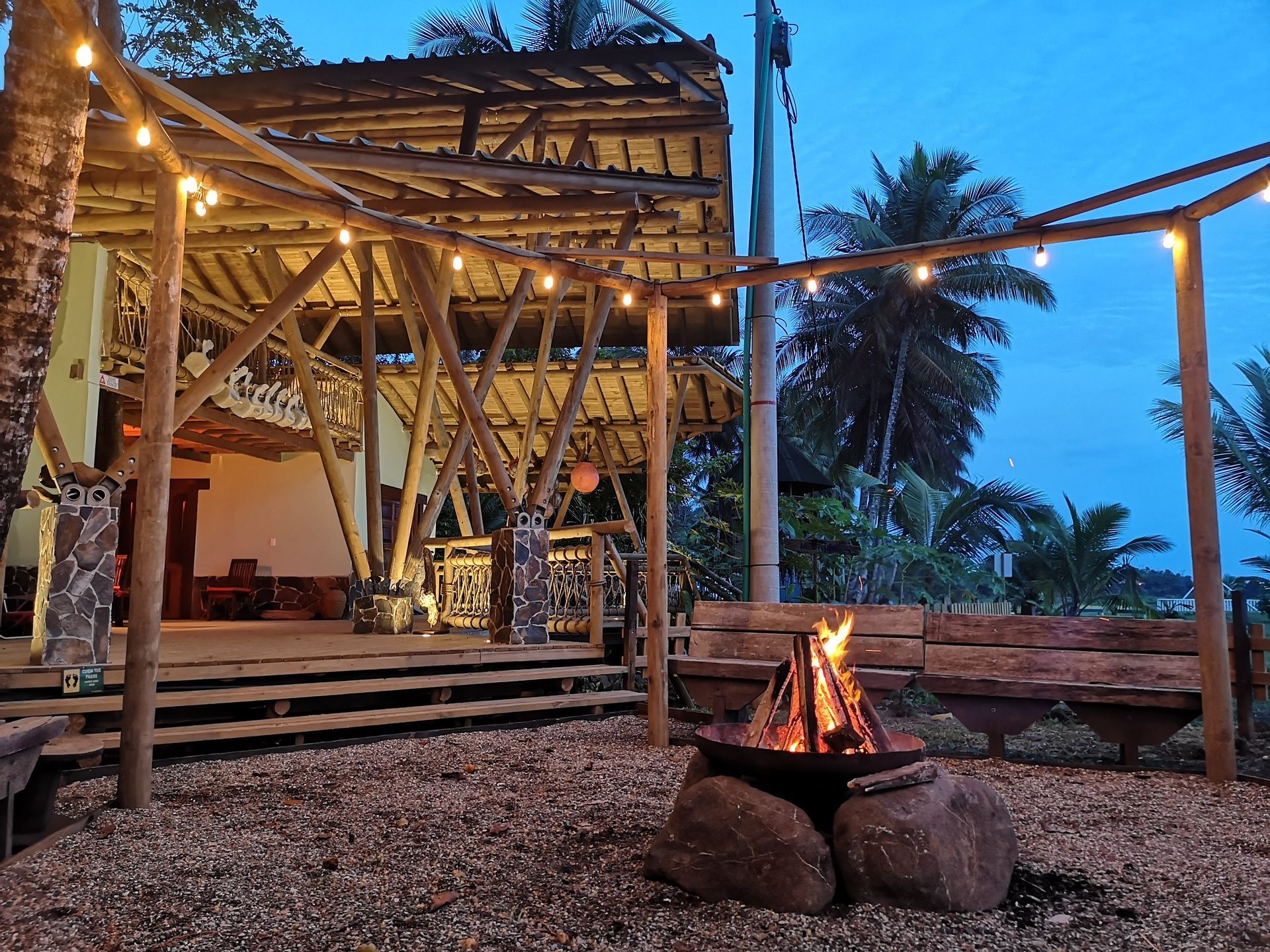 Tropical open-air patio at dusk with a lit fire pit, string lights, and wooden lounge seating