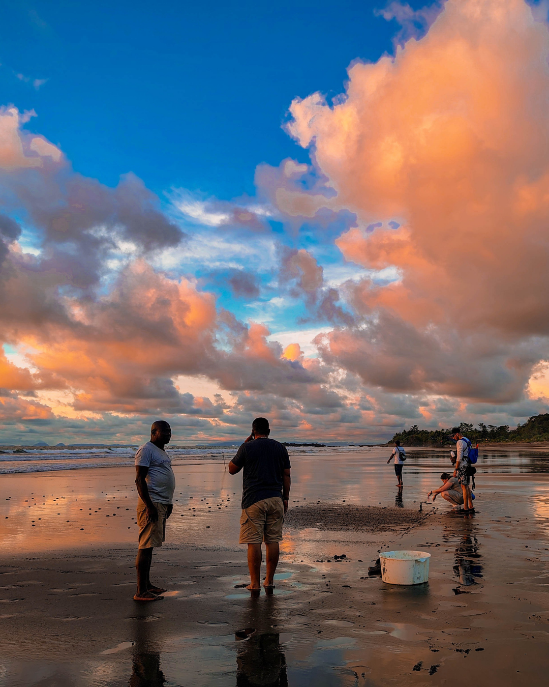 People standing on a wet beach at sunset under orange and blue clouds.