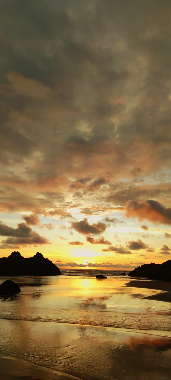 Golden sunset over a calm beach with reflective wet sand, dark clouds, and silhouetted islands on the horizon