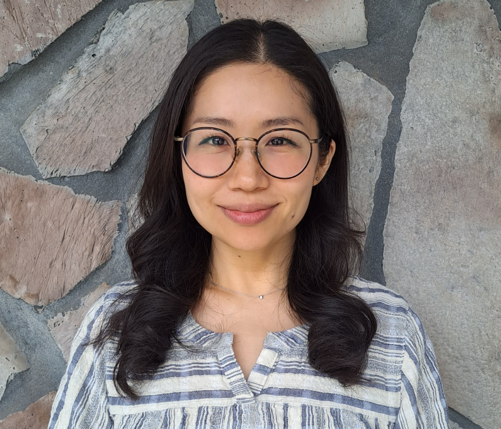 A woman wearing glasses and a striped shirt is standing in front of a stone wall.