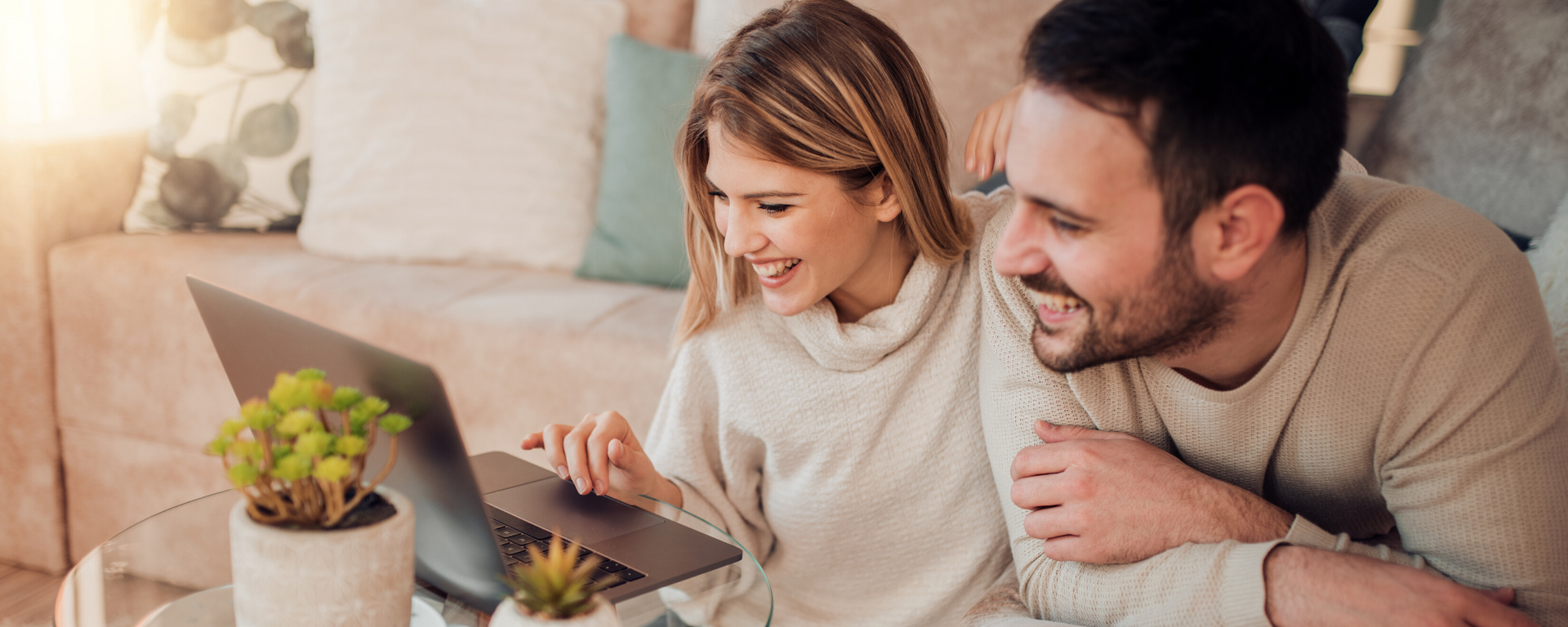 Two people smiling on a couch, looking at a laptop in a cozy living room