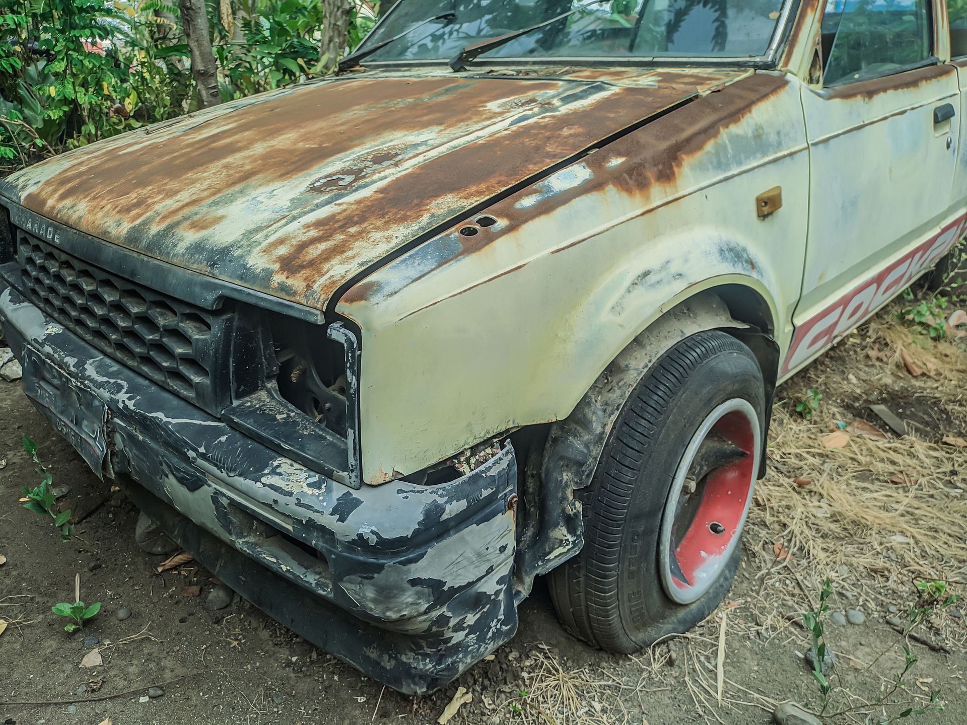 An old rusty car with a flat tire is parked in the dirt.