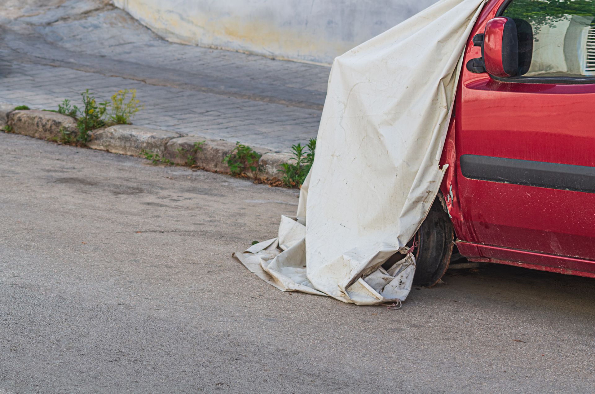 A red car is covered in a white tarp on the side of the road.