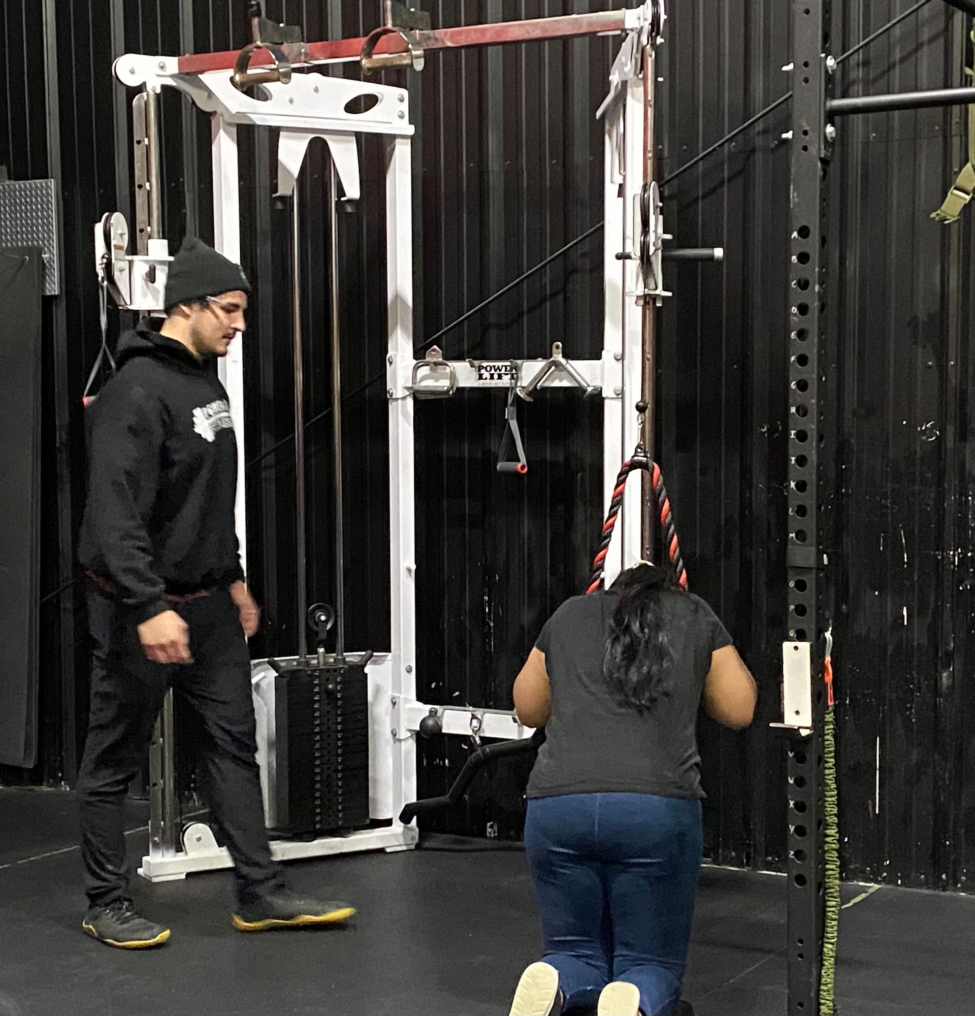 Man watches woman kneeling at a cable machine, pulling down on a rope at Power and Fitness gym in Bloomington, IL.