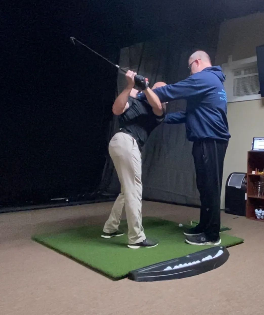 Golfer receiving instruction on a green mat indoors. Instructor touches golfer's shoulders, both are focused.