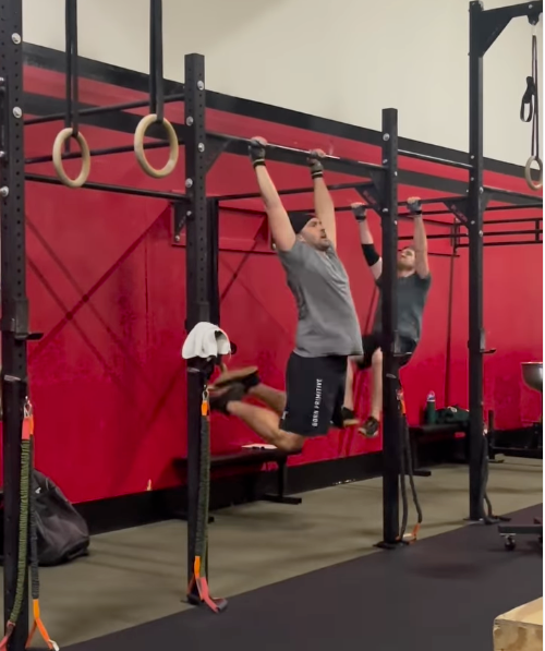 Two people doing pull-ups in a gym. One person has legs extended; both hang from a black pull-up bar with red walls.