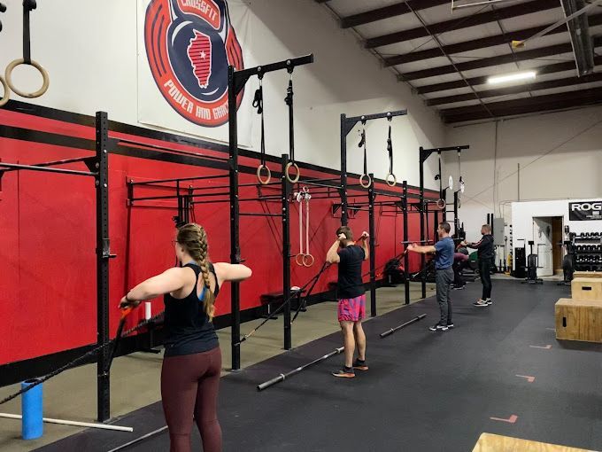 People working out with resistance bands in a CrossFit gym in Bloomington, IL. Red walls, black equipment, and a logo are visible.