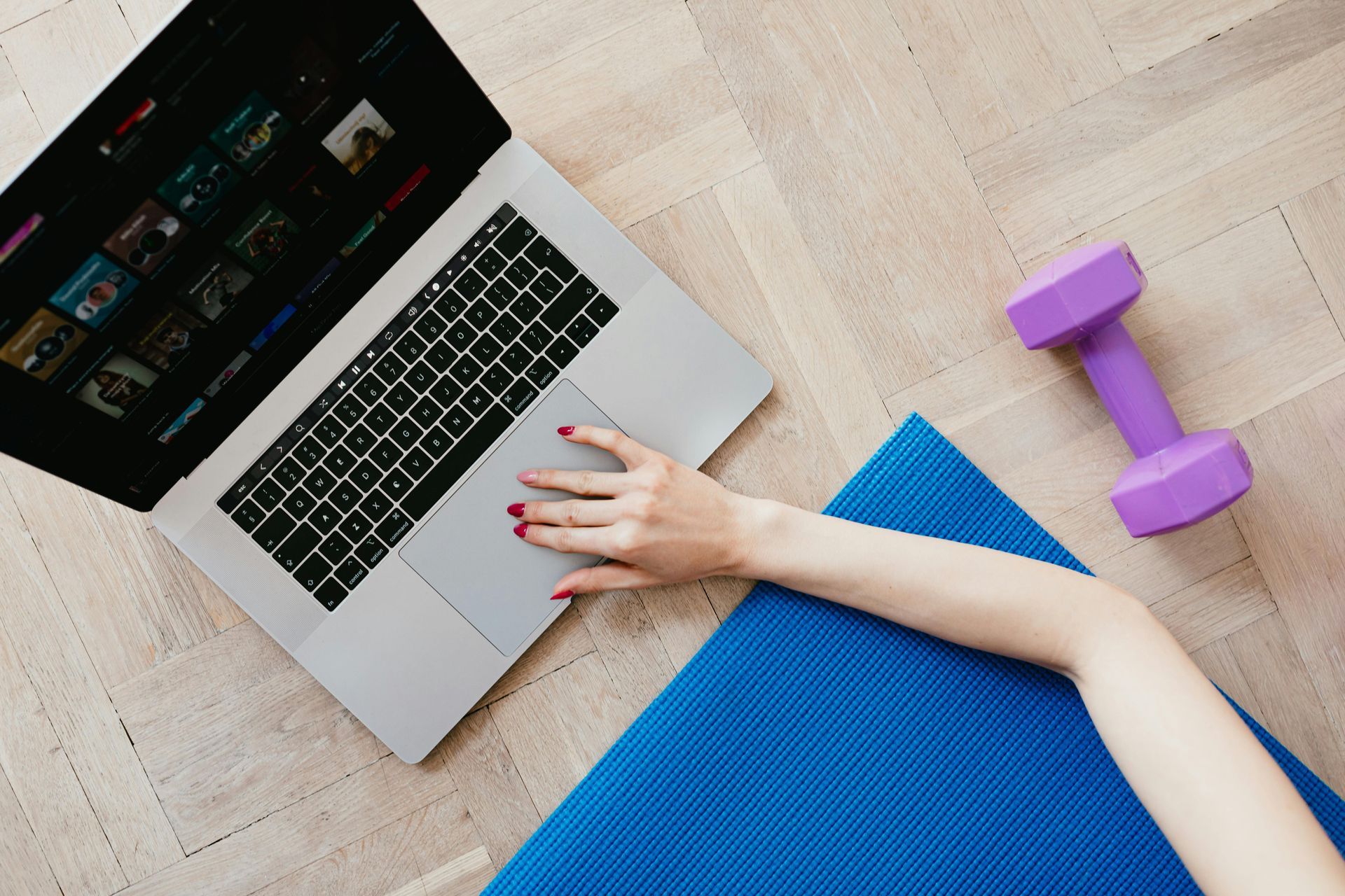 Woman's arm on laptop near blue yoga mat and purple dumbbell on wooden floor.