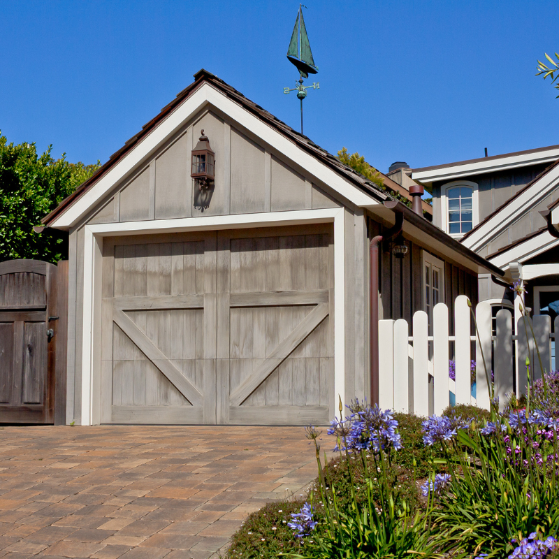 Wooden garage with weathered door, brick driveway, white picket fence, blue flowers, sunny day.