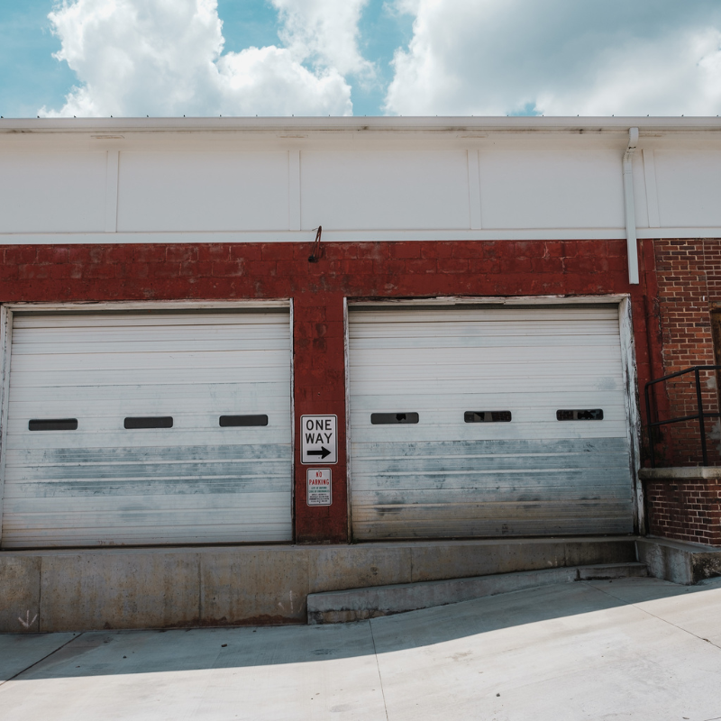 Two white garage doors on a red brick building, with a one-way sign. Concrete ramp leading up to doors.