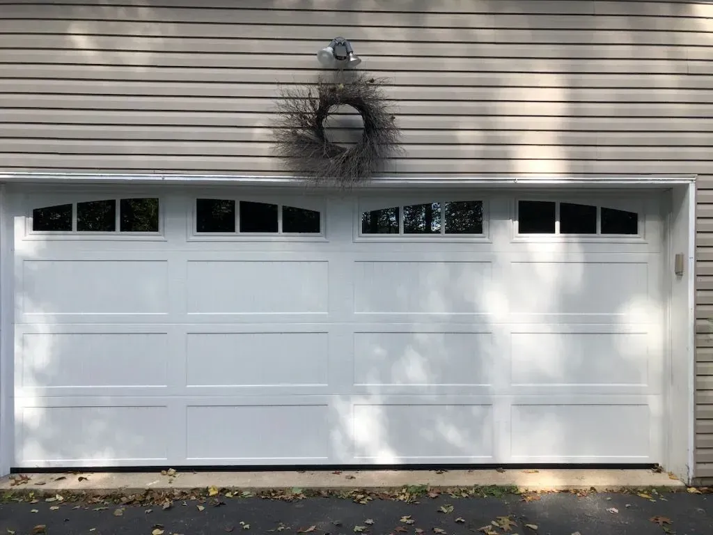 White garage door with windows, a wreath, and a light fixture against gray siding.