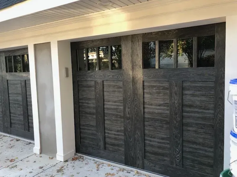 Gray wood-grain garage doors with rectangular glass panels, set in a white-trimmed doorway.