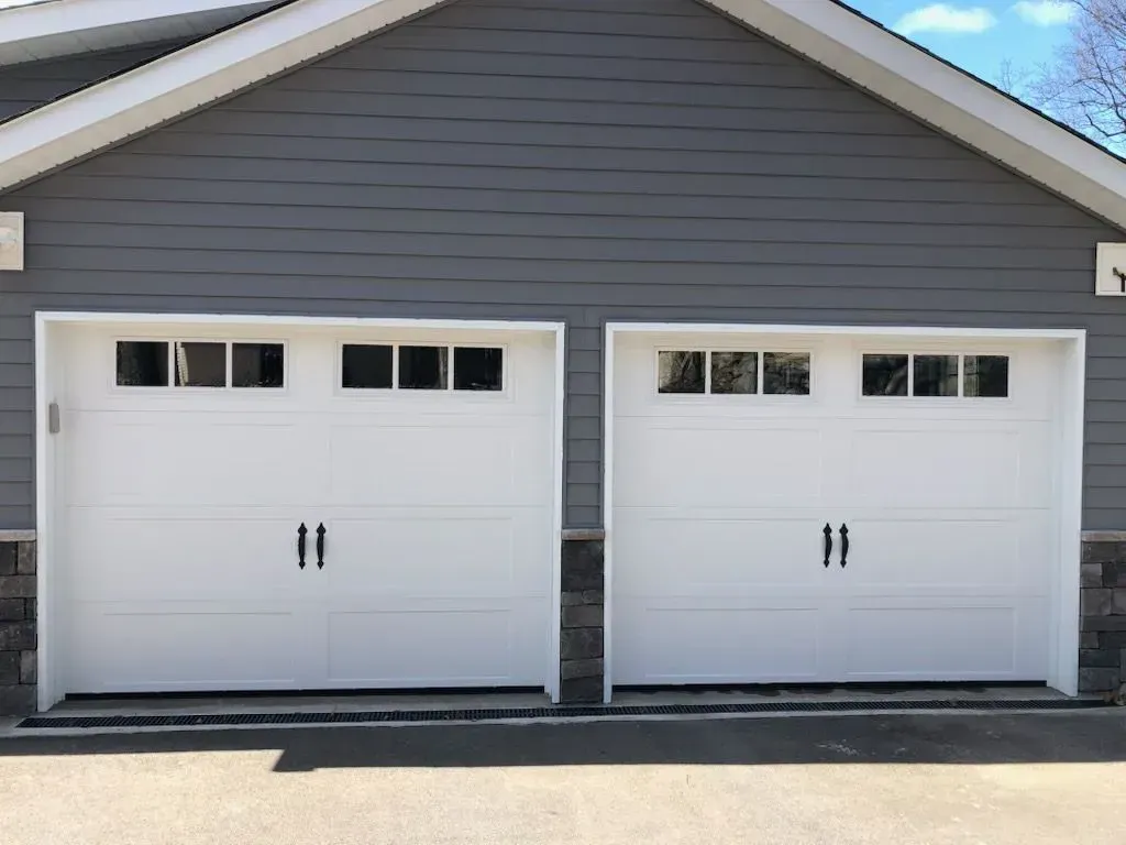 Two white garage doors with black handles, set in a gray-sided building with stone accents.