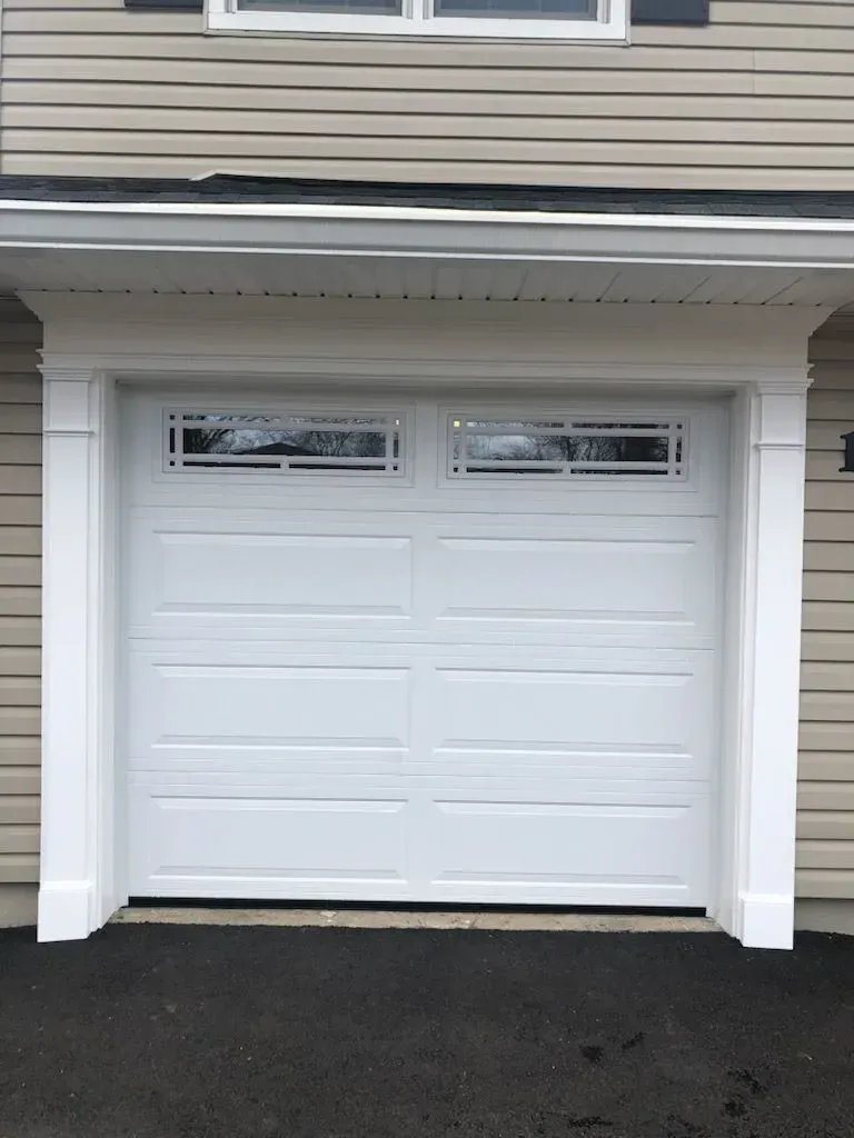 White garage door with glass panes, framed by white molding, against beige siding.