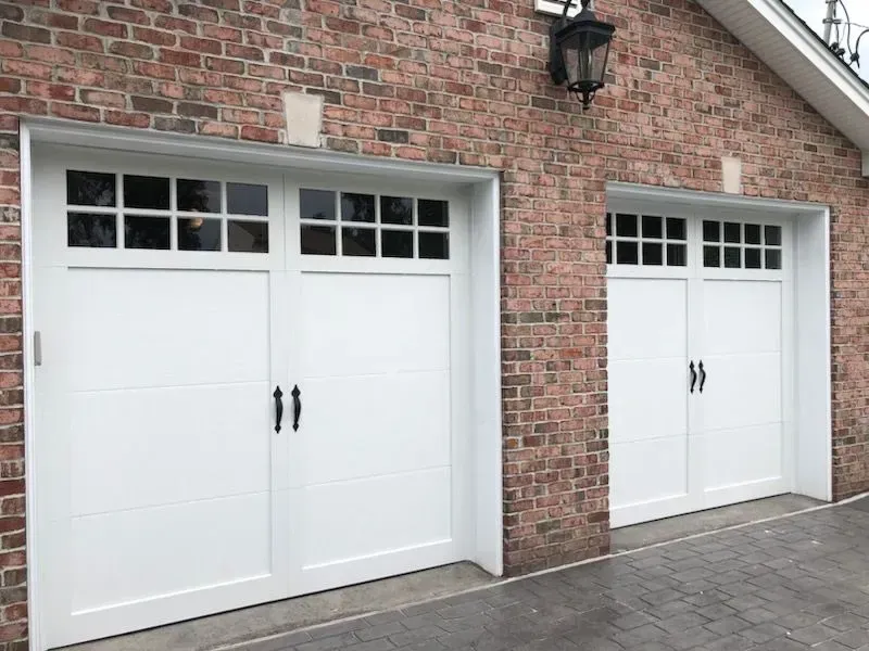 Two white garage doors with black handles and windows, set in a brick building.