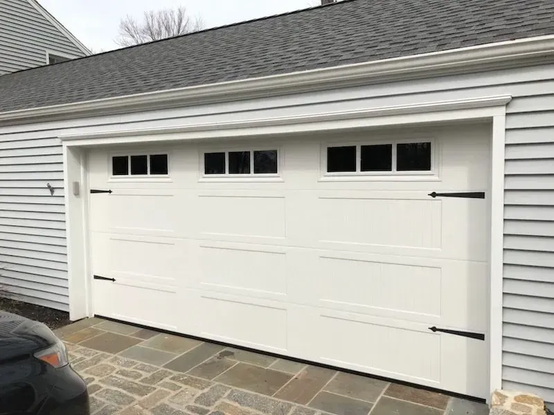 White garage door with windows, black hinges, and trim on a gray house.