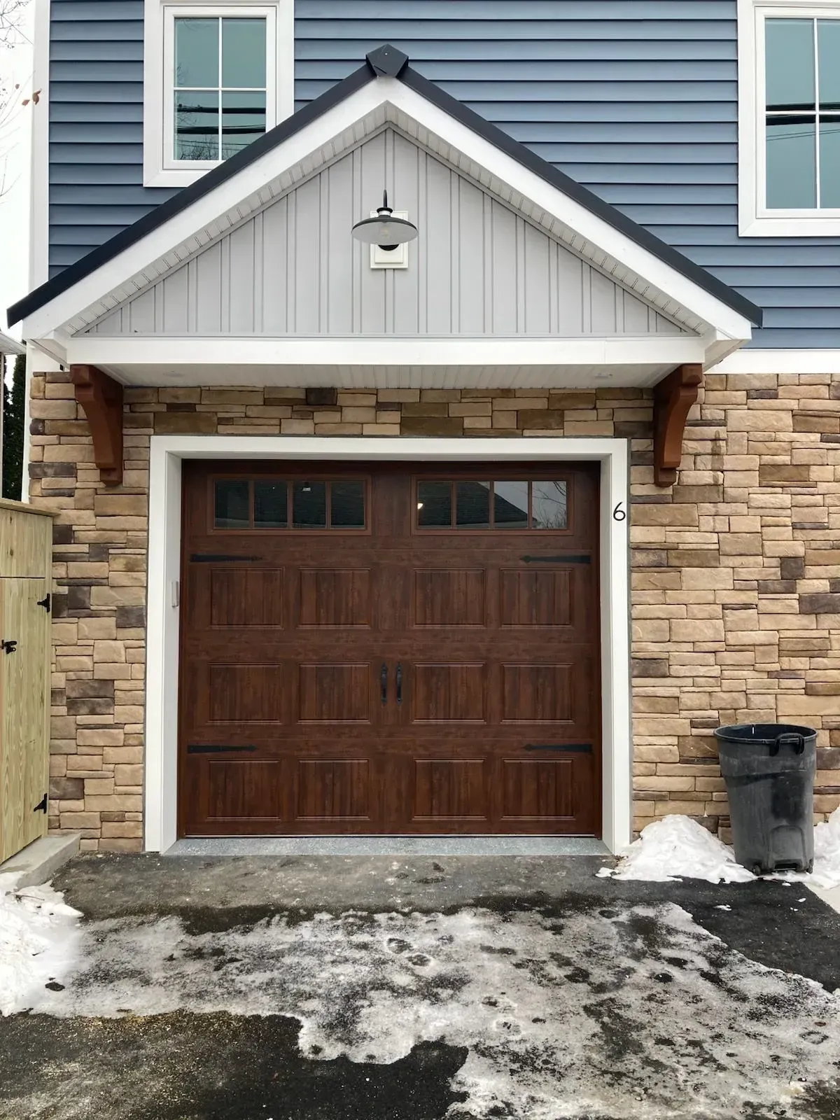Brown garage door under a white and gray gable, flanked by stone and blue siding, with a trash can and snow.