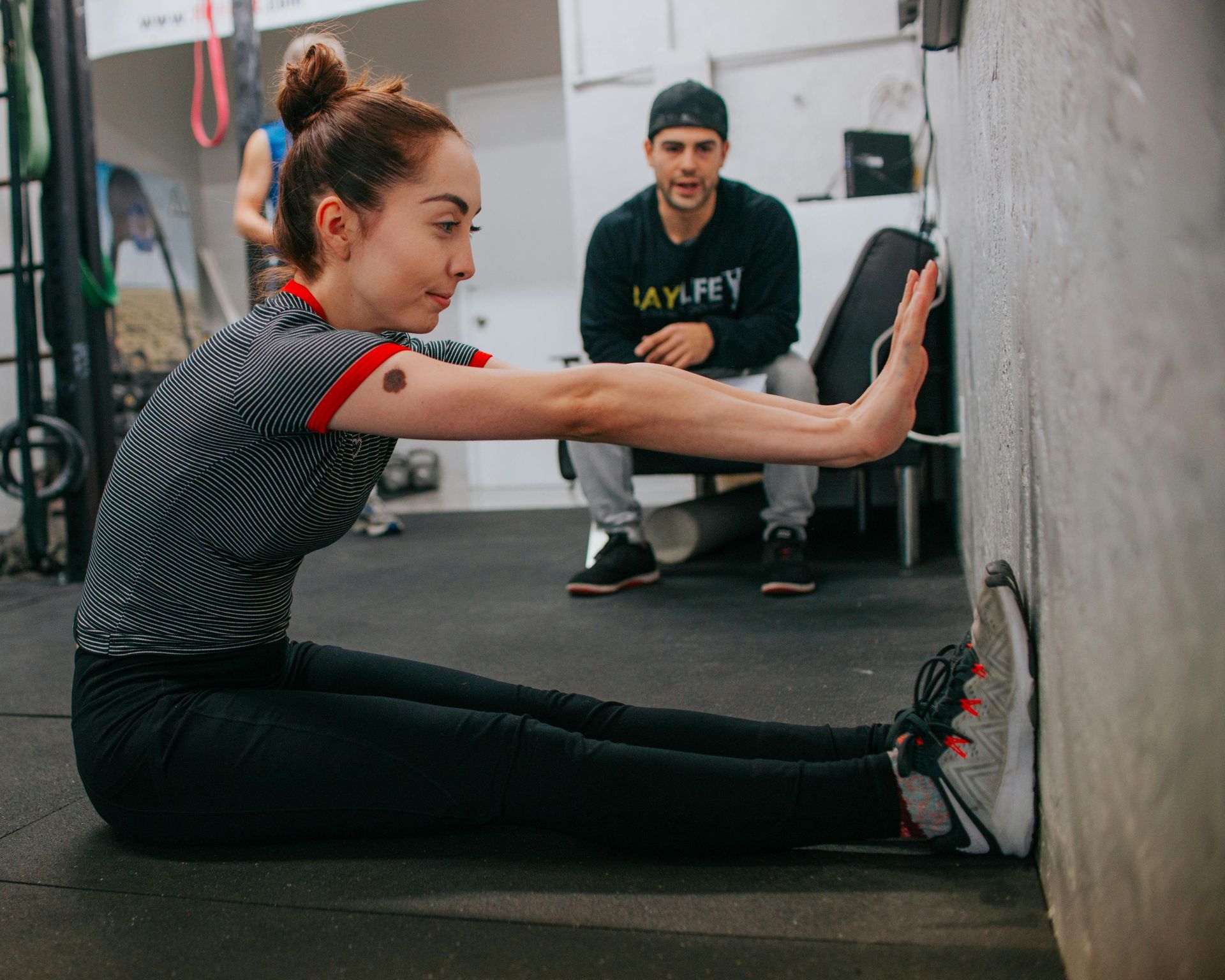 A woman is stretching her legs in a gym while her trainer watches.