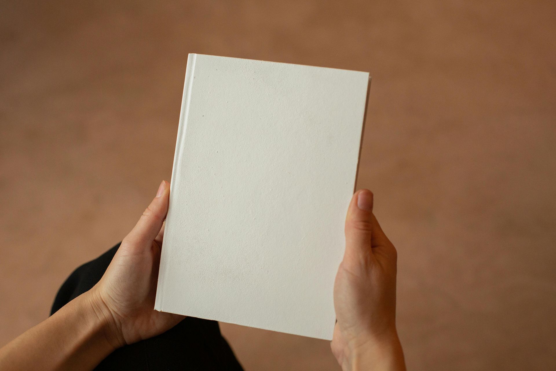 Hands holding a blank white book against a brown background.