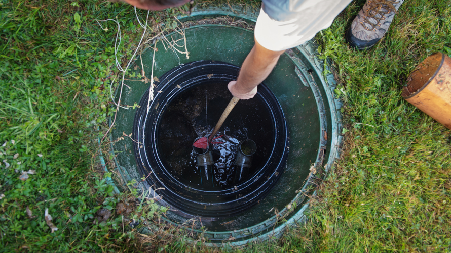 Person using a tool inside an open septic tank in a grassy yard.