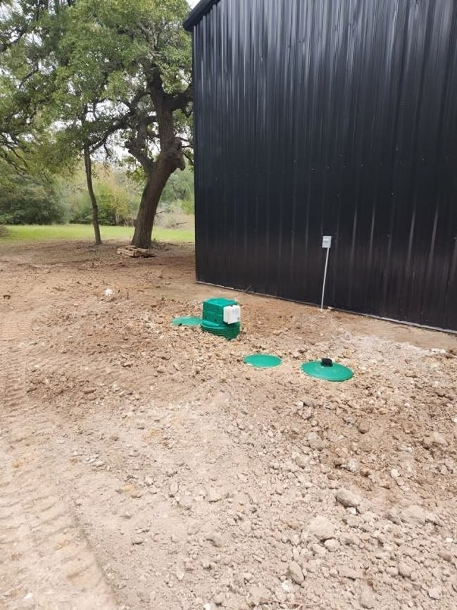 Green septic system components near a black building on a dirt lot, with a tree in the background.