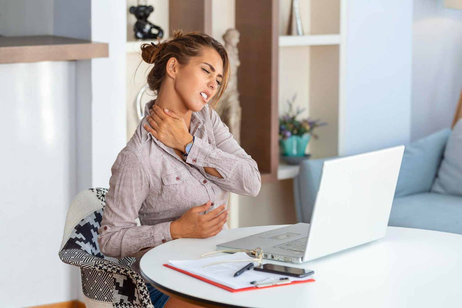 A woman is sitting at a table with a laptop and holding her neck.