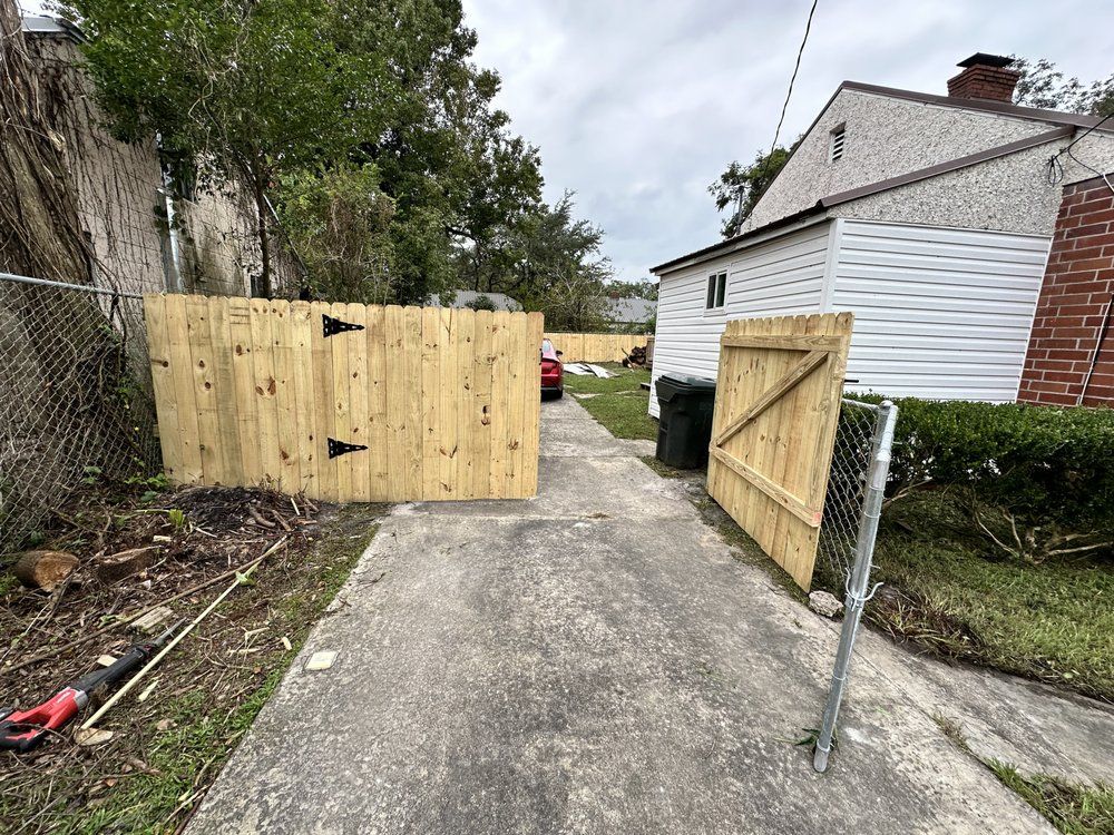 Wooden gate and fence enclose a concrete driveway leading to a backyard; a house is in the background.