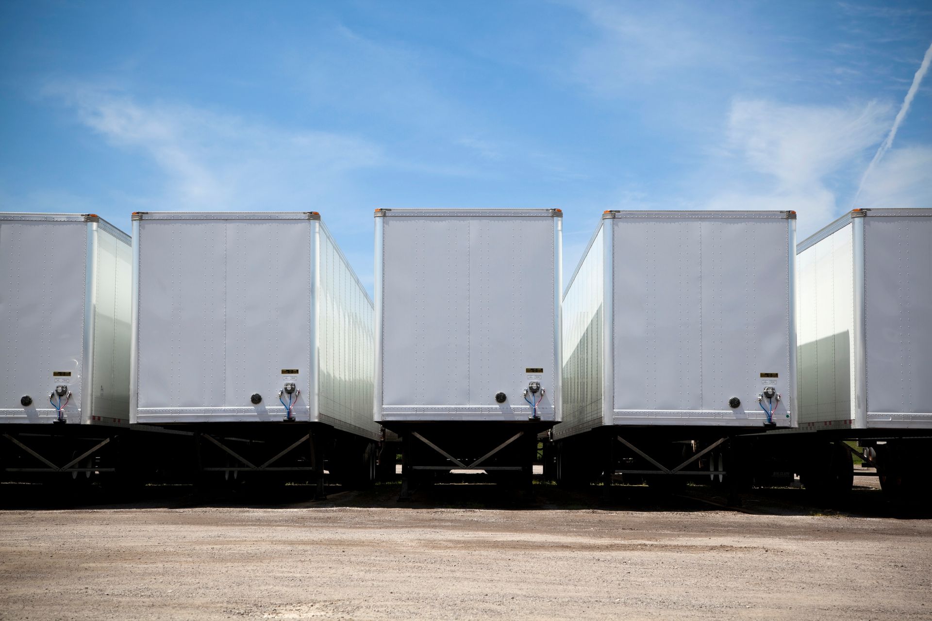 Row of white semi-trailer trucks parked on a gravel lot under a blue sky.