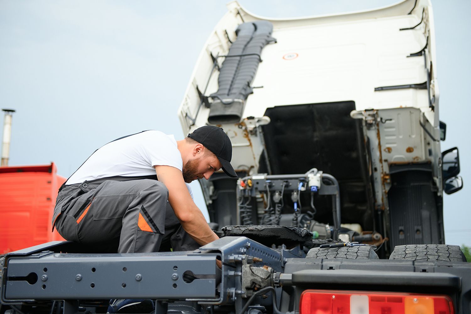 A mechanic wearing a black cap and white t-shirt is repairing a truck.