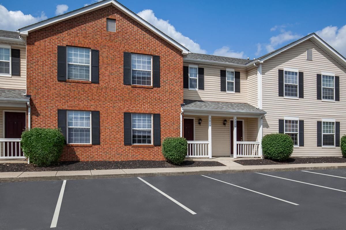 Row of townhouses, brick and beige siding, with black shutters and small porches, in a parking lot.