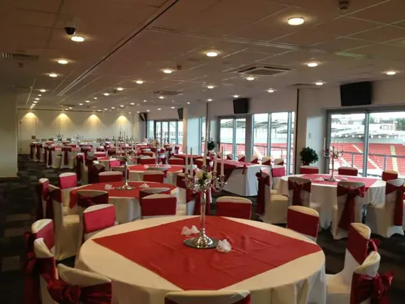 Red-and-white banquet hall with round tables, covered chairs, and large windows overlooking a waterfront.