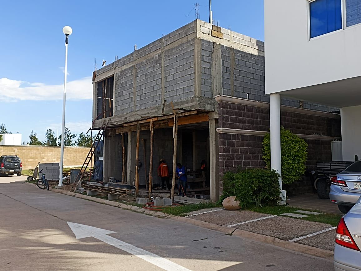 Edificio en construcción, paredes de bloques de hormigón gris, vigas de soporte de madera, cielo azul, dos trabajadores.