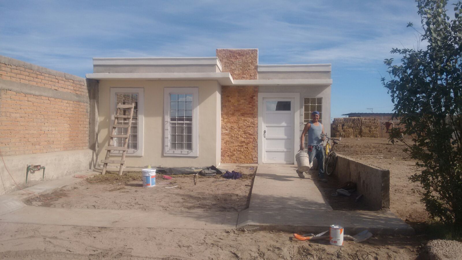 Pequeña casa en construcción con trabajador, escalera y suministros en un día soleado.