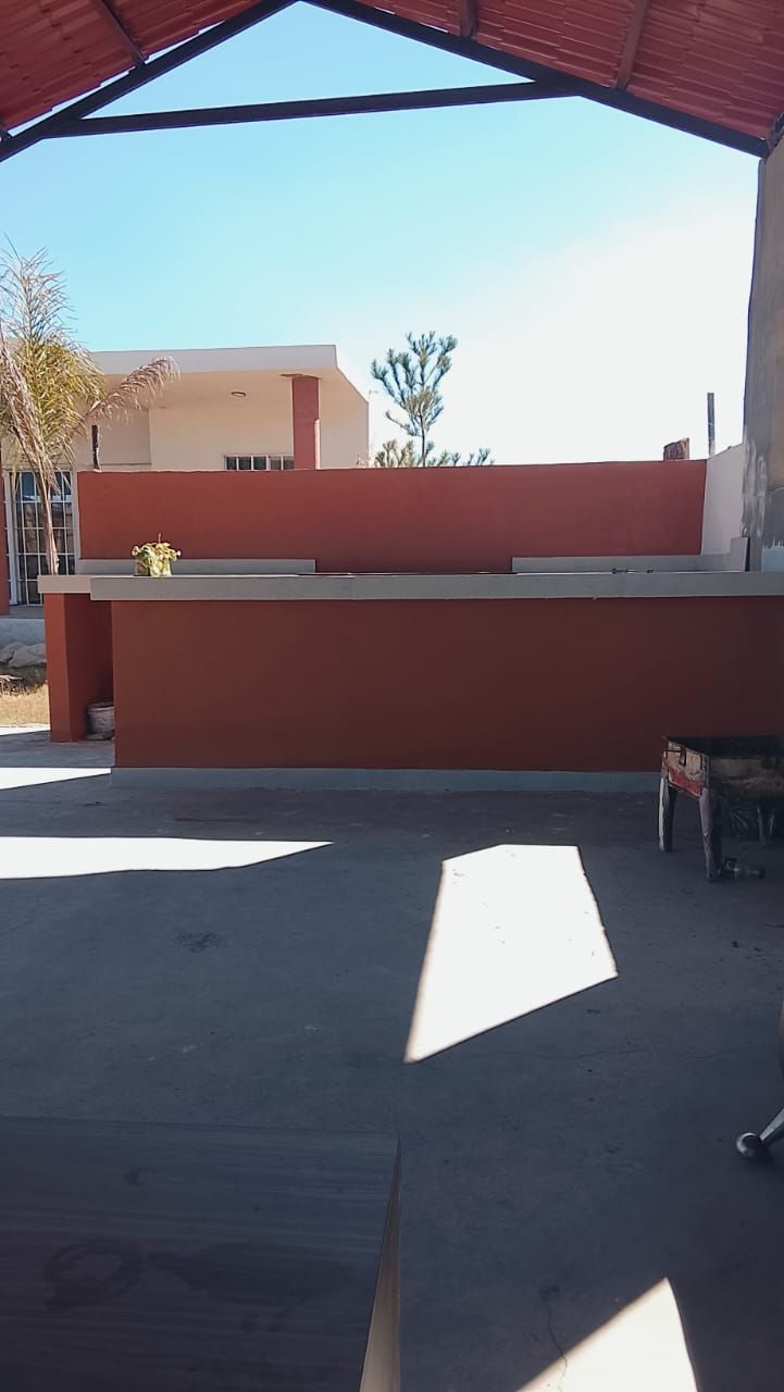 Cocina al aire libre con paredes rojas y encimera de hormigón bajo un área cubierta, fondo de cielo azul.