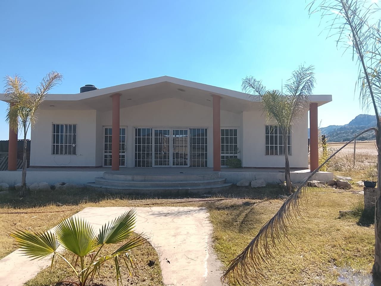 Un edificio blanco con techo inclinado y puertas de vidrio, ubicado en una zona verde bajo un cielo azul.
