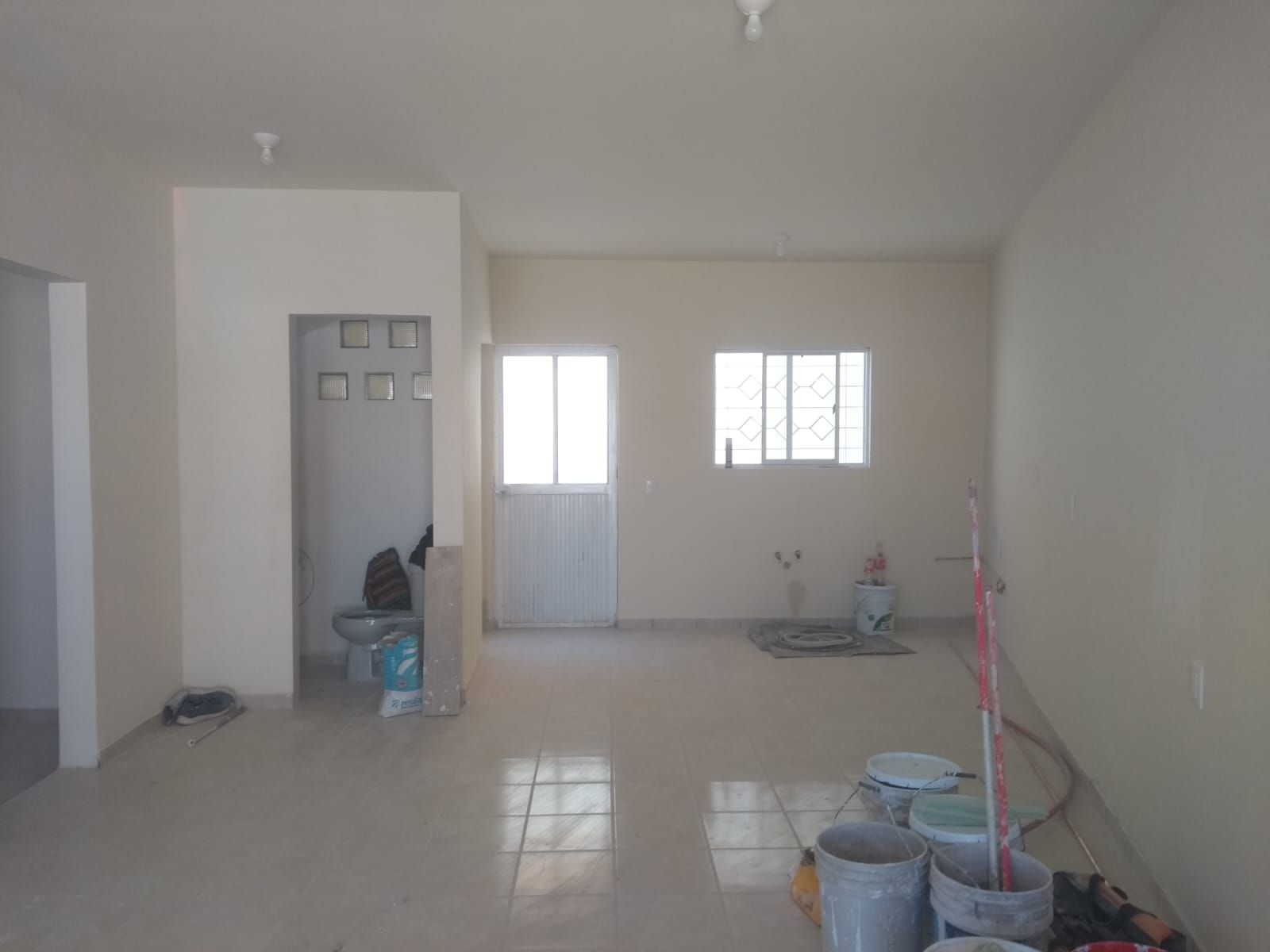 Habitación vacía con paredes y suelo de color claro. Hay puertas, ventanas y materiales de construcción.