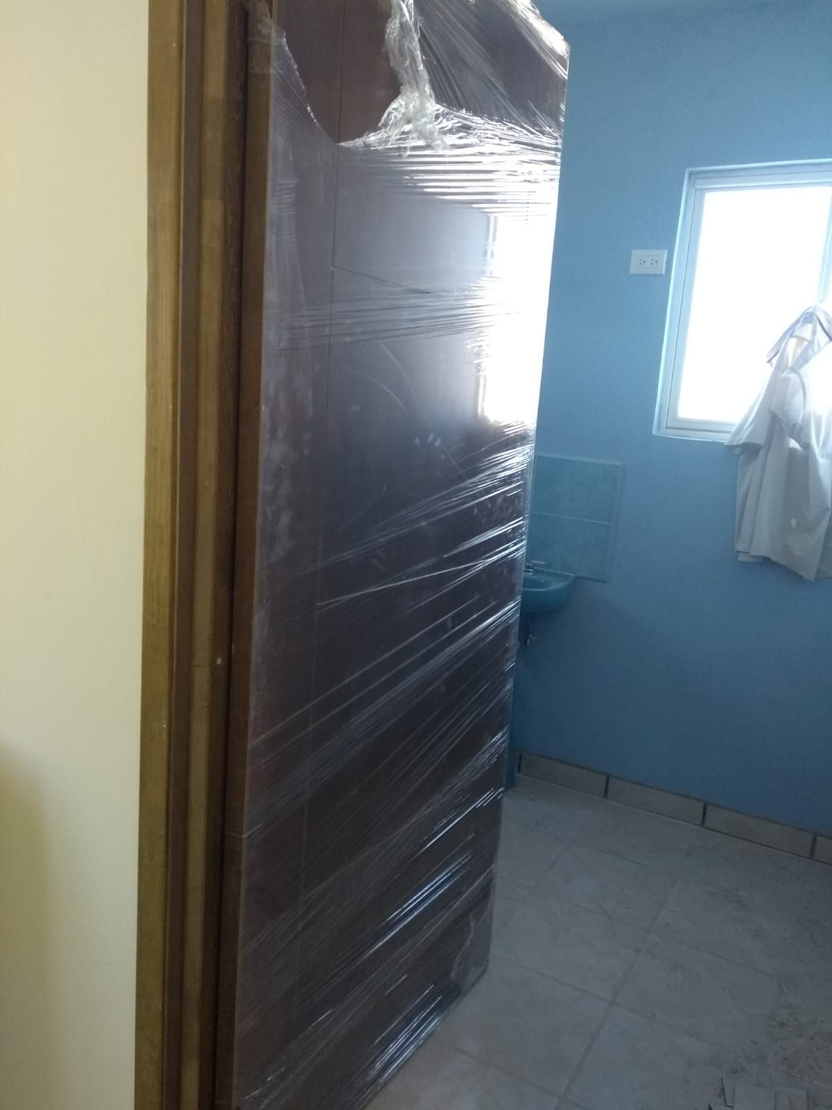 Puerta envuelta en plástico, vista a través de una puerta. Se ve una habitación azul con ventana y lavabo.