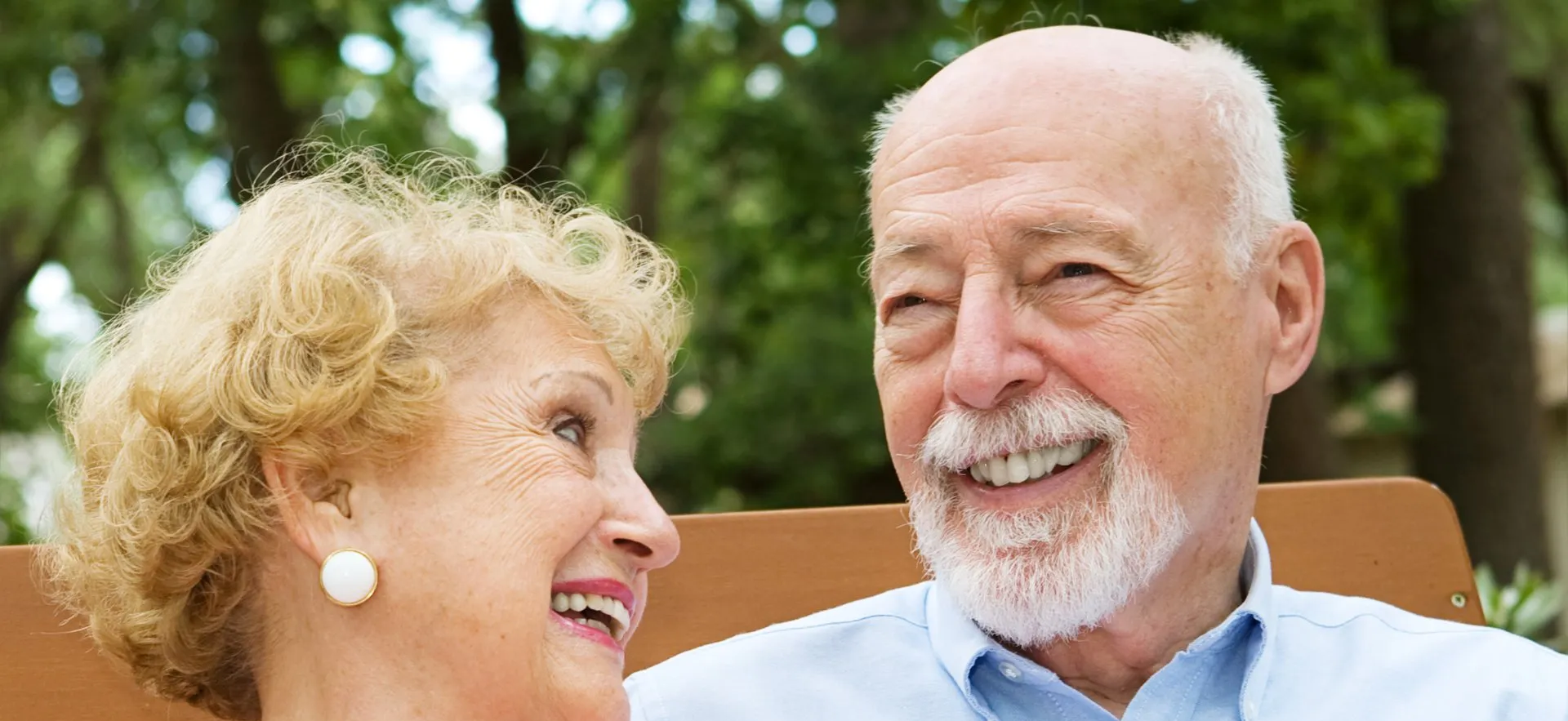 An elderly couple is sitting on a bench and smiling at each other.