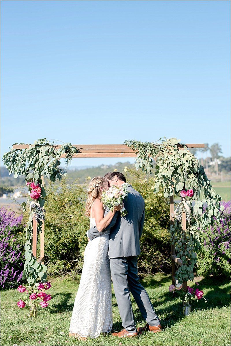 A bride and groom are kissing under a wooden arch at their wedding ceremony.